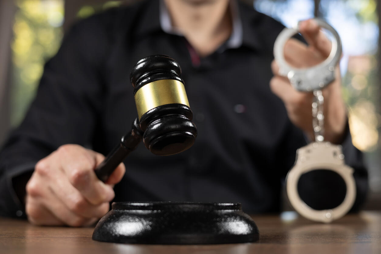 Close-up of a hand holding a gavel with handcuffs in the background, representing criminal justice and legal defense.